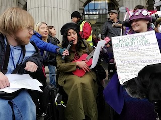 Protestors for Disability Power Ireland. 2023 A group of people participating in a protest, one person wearing a green coat playing a small guitar, another person holding a sign with protest messages, and a dog in the foreground