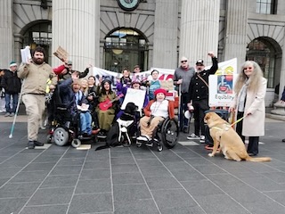 Disability Power Ireland Protest Dublin 2023 Group of people gathered outside a building with columns, some in wheelchairs, holding signs advocating for equality, accompanied by a service dog