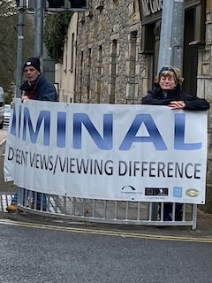 Liminal Different Views / Viewing Difference in Manorhamilton Two people holding a banner with the text 'LIMINAL Different views/viewing difference' on a street near a stone wall
